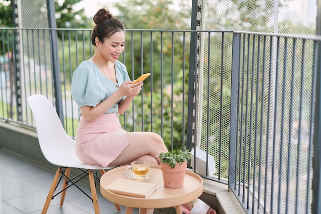 Asian young woman enjoying cup of coffee while using phone on the balcony