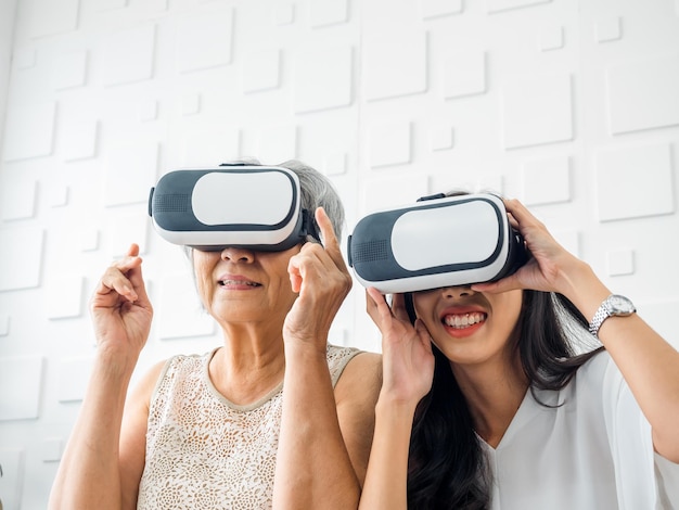 Asian young woman happy daughter and her mother old senior female wearing VR glasses enjoy 3d game online together in white room Mom and daughter with reality virtual technology lifestyle concept