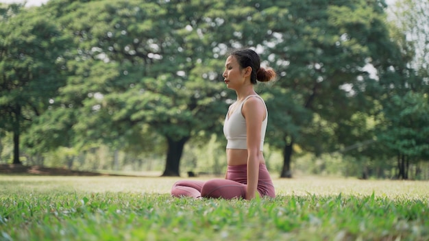 Asian young woman sitting on the grass in the lotus position and raising hands up outside in city park with the big trees background Rearview of female practicing yoga outdoors on a sunny day