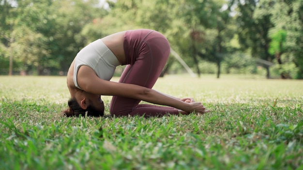 Asian young woman sitting on the grass practicing yoga in the cow pose position in city park with the big trees background Rearview of female practicing yoga outdoors on a sunny day