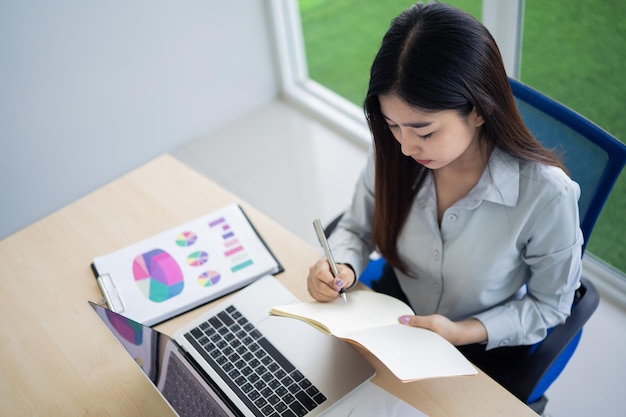 Asian young woman in smart casual wear writing on notebook and working on laptop while sitting in creative office or cafe young girl working with laptop on the wood table in the cafe