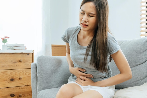 Asian young woman suffering stomach ache sitting on couch in living room at home people painful stomachache gynecology menstrual pain medical and health care concept