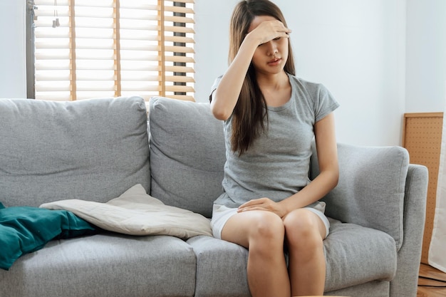 Asian young woman touching forehead having headache migraine and upset with problem feel stressed sitting on couch in living room at home people painful depression medical and health care concept