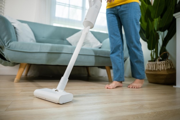 Asian young woman using vacuum cleaner dust while cleaning floor at home, homemaker easy cleaning with modern wireless vacuum cleaner near sofa in living room, housework concept