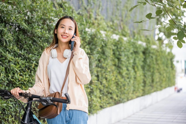 Asian young woman walking in countryside outdoor with her bicycle she using mobile phone talking with friend, Happy female stand on city street with bike using smartphone for summer sightseeing walk