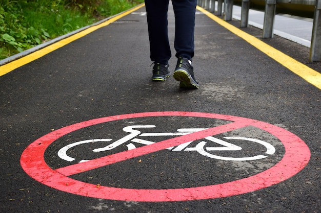 On the asphalt pedestrian sidewalk in the foreground, there is an imprint of a sign that is not allowed to travel to cyclists. In the background are the legs of a walking man in sneakers.