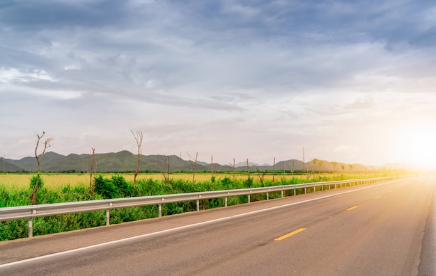 Asphalt road beside green grass field and the mountain with sunlight. Long distance journey with blue sky and white cloud. Country asphalt road. Road trip travel concept.