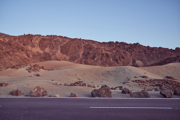 Asphalt roadway running along rough rocky mountains located near sandy terrain against cloudless blue sky in nature on summer day