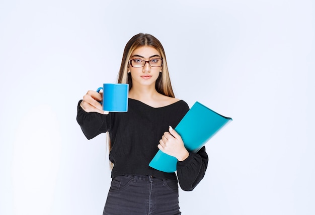 Assistant with eyeglasses holding a blue folder and offering a blue cup of drink to her colleague. High quality photo