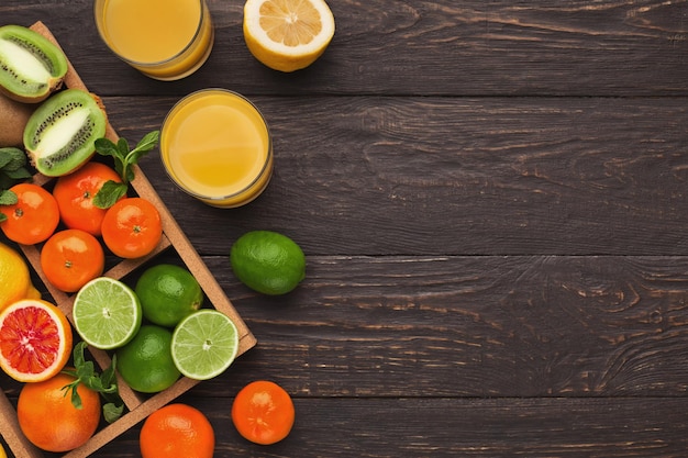 Assorted citrus fruits and fresh juice glasses for breakfast on dark wooden background. Top view on box with oranges, lemons, tangerines and other exotic fruits, flat lay, copy space