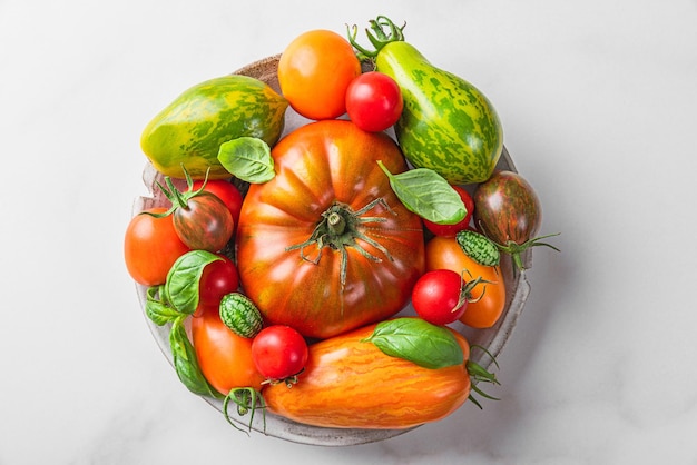 Assorted colorful organic tomatoes with mini cucumbers and basil in a plate on white table Top view