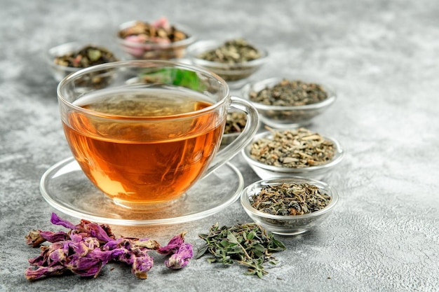 Assorted dried herbs with cup of tea in glass bowls on plaster surface