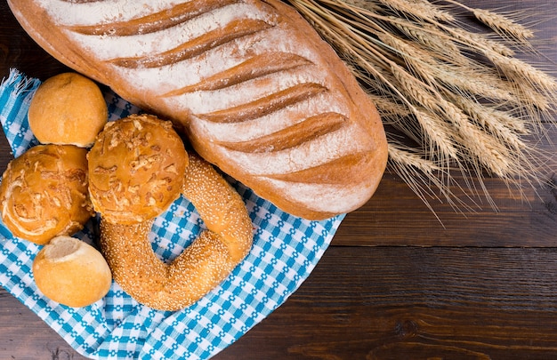 Assorted fresh bread rolls and baguette with ears of wheat displayed on a colorful blue and white cloth on a wooden table, overhead view