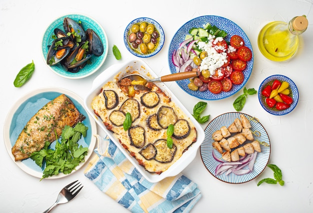 Assorted Greek dishes on white stone background from above, moussaka, grilled fish, souvlaki, greek salad, steamed mussels with herbs, appetizers of Greece from above