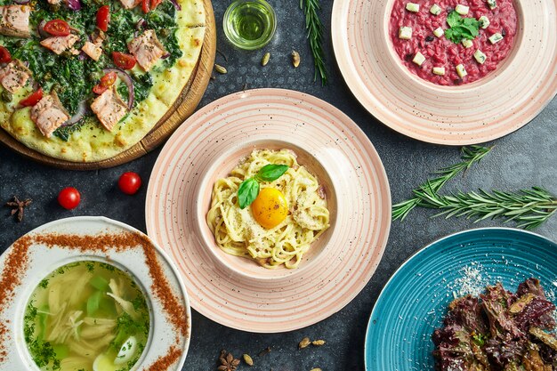 Assorted Italian food on a dark surface: carbonara pasta, salmon pizza, beetroot risotto, homemade chicken broth and salad. Top view flat lay food