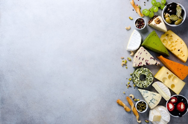 Assortment of hard, semi-soft and soft cheeses with olives, grissini bread sticks, capers, grape, on grey concrete. 