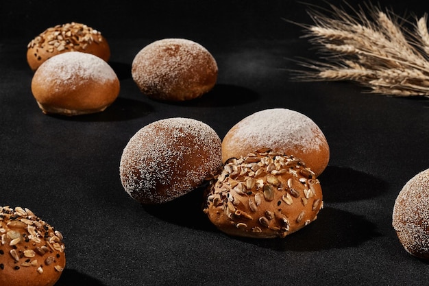 Photo assortment of tasty baked buns with sesame and sunflower seeds sprinkled with flour bunch of wheat against black background copy space closeup