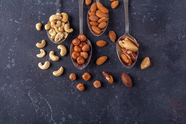 Assortment of various types of nuts: cashew, hazelnuts, almonds, brazil nuts on metal silver spoons on dark background.