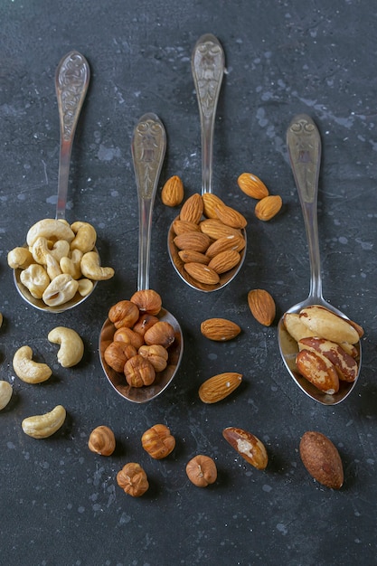 Assortment of various types of nuts: cashew, hazelnuts, almonds, brazil nuts on metal silver spoons on dark background