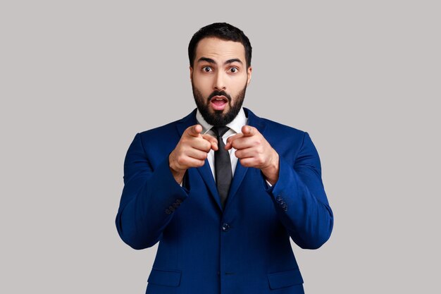 Astonished bearded man pointing to camera, standing with surprised shocked expression on face, hard to believe, wearing official style suit. Indoor studio shot isolated on gray background.