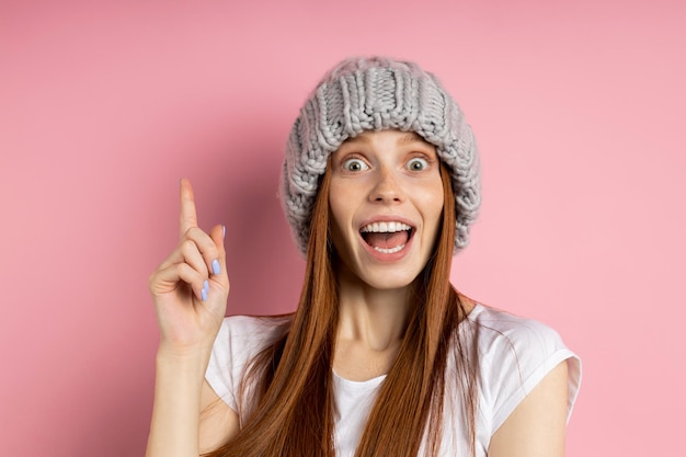 Astonished female with long ginger hair, freckles pointing upwards with index looking at camera with eyes popped out and wide opened mouth. People, emotions and promotion concept.