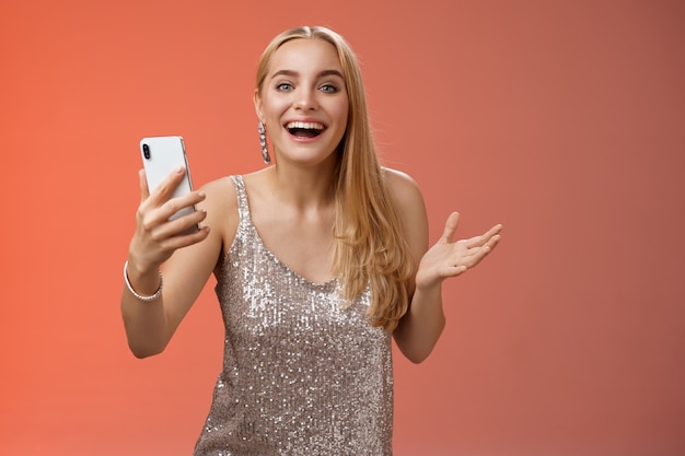 Astonished happy charming blond girl in silver glittering stylish dress holding smartphone amazed liking awesome result edit photo app smiling wondered amused, standing red background.