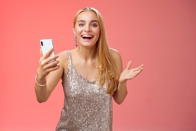 Astonished happy charming blond girl in silver glittering stylish dress holding smartphone amazed liking awesome result edit photo app smiling wondered amused, standing red background.