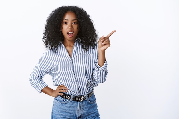 Astonished impressed woman asking friend what was that pointing sideways questioned curious indicating side index finger open mouth intrigued standing casually hand waist, wearing striped blouse