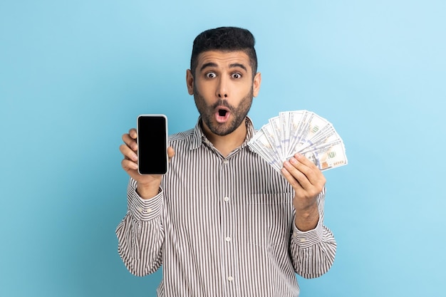 Astonished man with beard holding cell phone and dollars banknotes shocked by money cashback