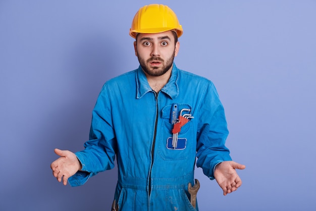 Astonished shocked young builder wearing yellow helmet and blue uniform, standing with opened mouth and spreading hands