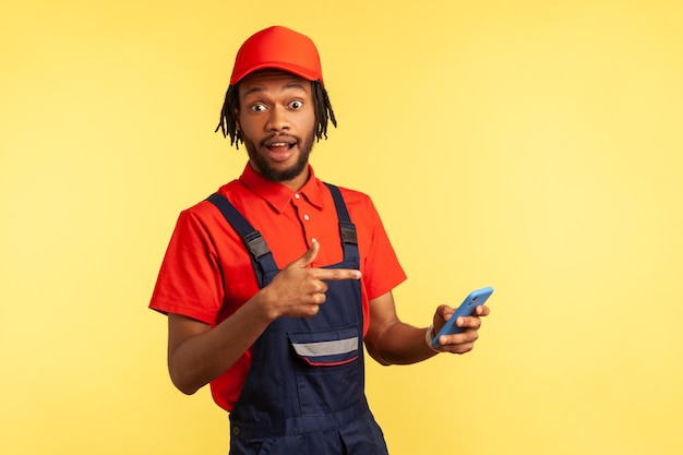 Astonished worker with beard wearing uniform pointing at mobile phone with shocked expression, profitable proposition in delivery service. Indoor studio shot isolated on yellow background.