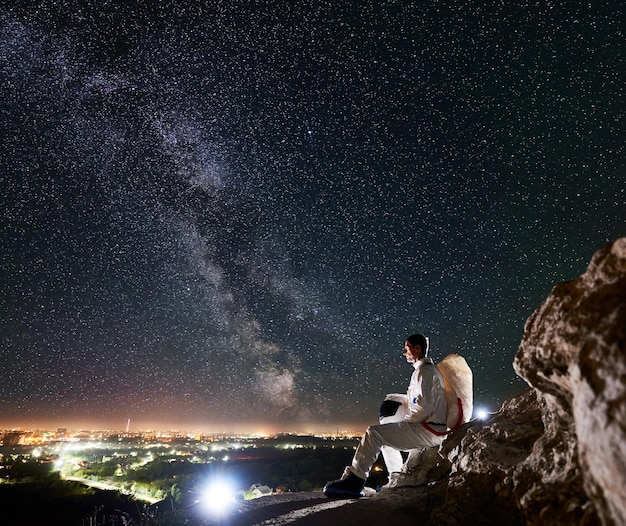 Astronaut sitting on rocky hill under beautiful night sky with stars