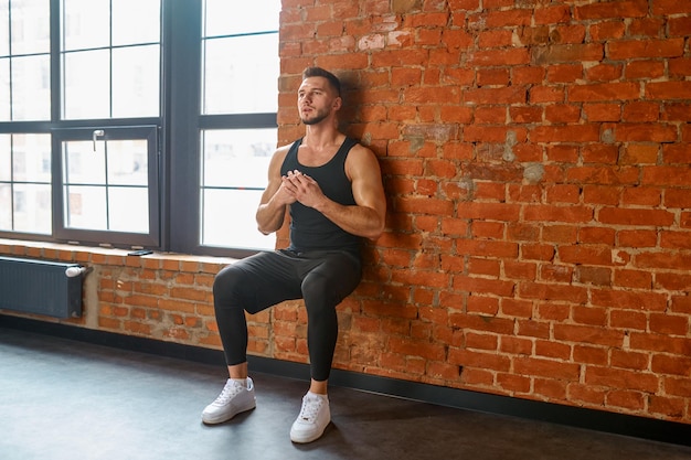 Athlete doing training exercise against gym wall. Sportsman exercising squats leaning back on brickwall. Sport, bodybuilding, fitness and people concept