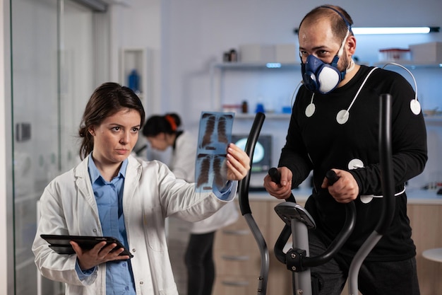 Athlete man with electrodes sensors on it running on fitness bycle working at body endurance in medical laboratory while specialist researcher analyzing bones radiography. Doctor monitoring EKG data