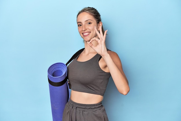 Athlete with a mat in blue studio cheerful and confident showing ok gesture