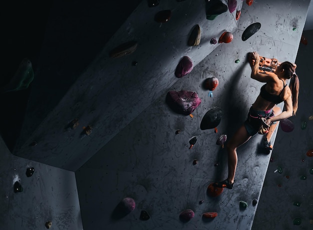 Athlete young woman in shorts and sports bra exercising on a bouldering wall indoors.