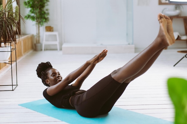 Athletic afro american woman lying on back and doing fold exercise with outstretched legs and arms on gym mat in photo studio Sports course exercise technique pumping up press beautiful figure