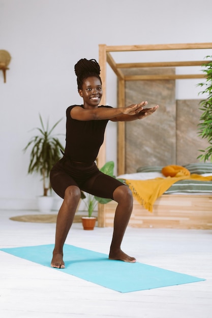 Athletic afro american woman with smile squatting with arms extended forward training on gymnastic mat in decorated studio Muscle pumping sports exercises fitness keeping body fit