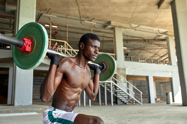 Athletic black young man lifting a heavyweight barbell in outdoor gym under the bridge Healthy lifestyle concept