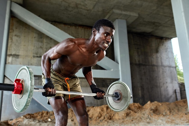 Athletic black young man lifting a heavyweight barbell in outdoor gym under the bridge Healthy lifestyle concept