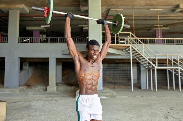 Athletic black young man lifting a heavyweight barbell in outdoor gym under the bridge healthy