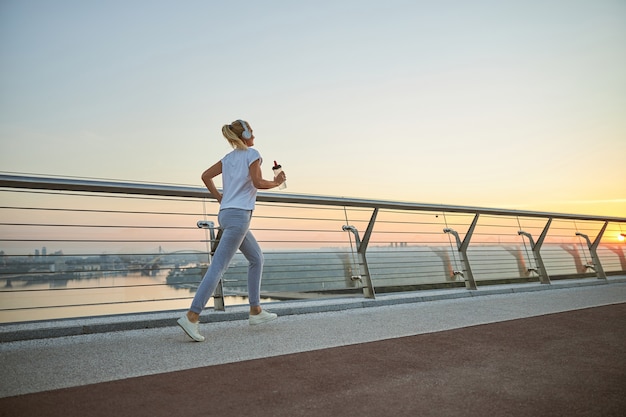 Athletic blonde female in headphones and with a bottle of water jogging across the footbridge