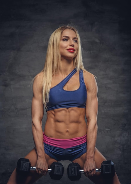 Athletic blonde fitness model posing with dumbbells on grey background.