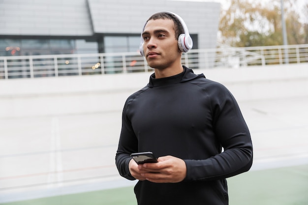 Photo athletic caucasian sportsman in tracksuit using headphones and cellphone while working out on stadium after rain