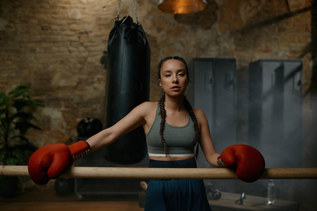 Athletic female fighter with red boxing gloves standing leaned at wooden training bar in gym with loft interior design