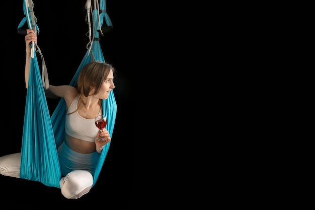 Athletic girl practices aerial gymnastics or fly yoga in hammock with glass of wine on black background Copy space