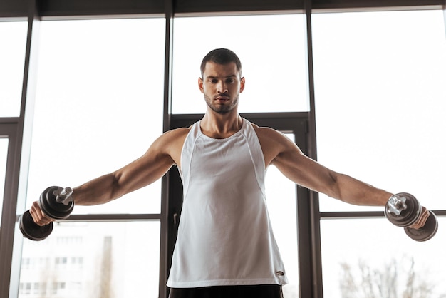 Athletic man with outstretched hands exercising with dumbbells in gym