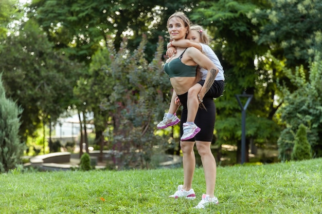 Athletic mom exercising with her little daughter on her back outdoors healthy lifestyle fitness active family concept Training together