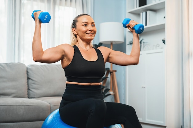 Athletic and sporty senior woman sitting on fit ball while engaging in weight lifting with dumbbell at home exercise as concept of healthy fit body lifestyle after retirement Clout