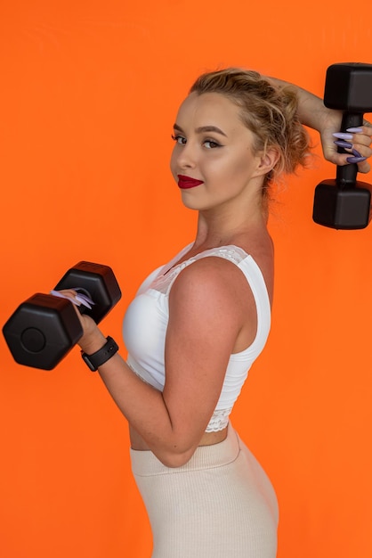 An athletic wellgroomed woman with cute appearance stands with dumbbells on plain background